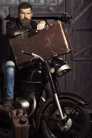 Handsome unshaven male biker in leather jacket sitting near motorcycle in garage with big brown old briefcase and rusty fuel can looking forward on workshop background, vertical pictureの写真素材