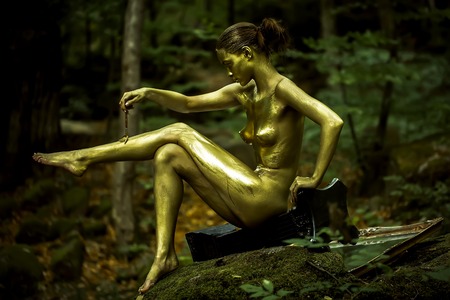 Beautiful young undressed woman with golden body painting legs with brush sitting on column in deep green forest on stona with moss outdoor on natural background, horizontal pictureの写真素材