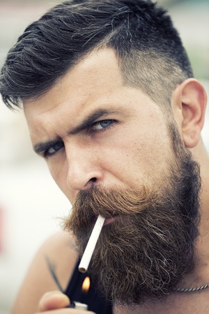 Portrait of sexual young unshaven man with beard and handlebar moustache smoking cigarette looking forward with expressive face outdoor closeup, vertical pictureの写真素材