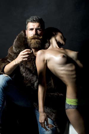 Young couple of undressed girl in panties standing near unshaven man with beard and moustache smoking cigarette in fur coat on black background, vertical pictureの写真素材