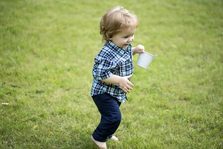 Cute small baby boy with blond curly hair in blue checkered shirt and jeans holding white bucket running on green grass lawn sunny day outdoor copyspace, horizontal pictureの写真素材