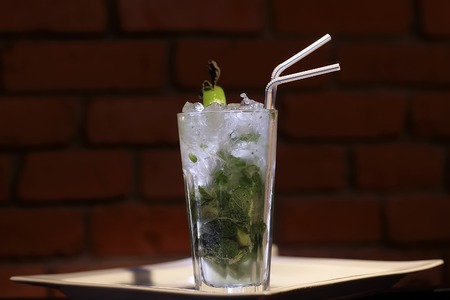 One glass goblet with alcoholic mojito cocktail of light cuban rum fresh lime juice fresh mint leaves granulated sugar soda water and ice cubes and drink straw on brick background, horizontal photoの写真素材