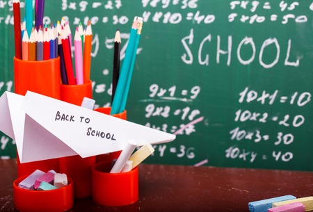 Colorful pencils of red yellow orange violet purple pink green and blue in stationary cup and paper plane with back to school text on desk on written with white chalk blackboard, horizontal photoの写真素材