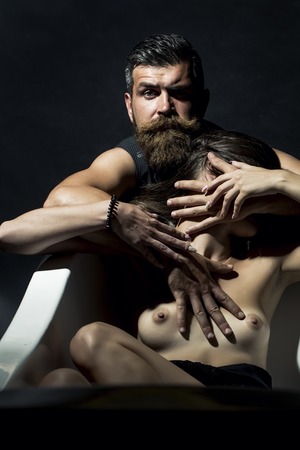 Young couple of sensory bare girl lying in white bathtub and unshaven man with beard and moustache in waistcoat sitting on black background, vertical pictureの写真素材