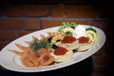 White round plate with many different kinds of seafood of red salmon caviar shrimp bread butter yellow lemon black olive berry lettuce and greenery on brown brick wall background, horizontal pictureの写真素材