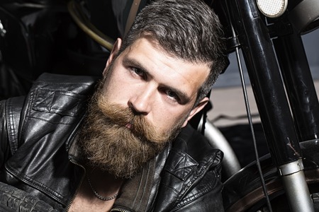 Portrait of handsome pensive unshaven man with beard and handlebar moustache in black leather biker jacket sitting near motorcycle looking forward, horizontal pictureの写真素材
