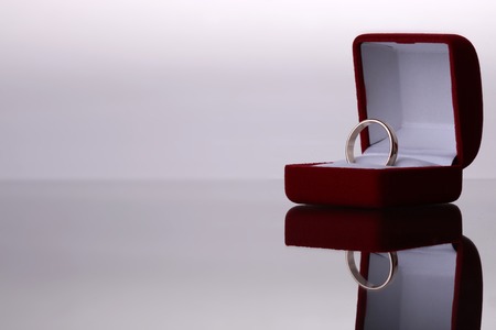 Closeup of one small beautiful velvet red open wedding box with white fabric inside with gold or silver engagement ring standing on grey reflecting table top on studio isolated background copyspace, horizontal pictureの写真素材
