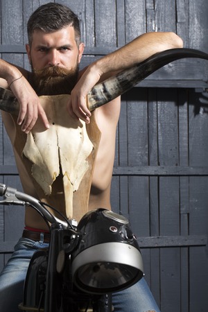 Handsome sexy undressed unshaven male biker in jeans sitting on motorcycle in garage with big bone skull antlers of stuffed animal looking forward on wooden wall , vertical pictureの写真素材