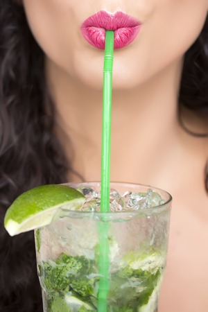 Closeup of sexual pretty brunette female face of girl with curly hair and pink lips drinking alcoholic mojito cocktail from mint soda light rum and lime with straw, vertical pictureの写真素材