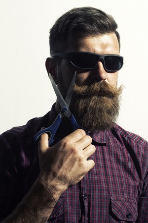 Portrait of unshaven man in violet checkered shirt and sunglasses with long beard and handlebar moustache holding scissors in hand standing isolated on white , vertical pictureの写真素材