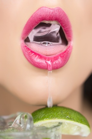 Closeup of sexy open female mouth of attractive young woman with bright pink lips holding cold fresh crystal ice cube from glass of alcoholic mojito cocktail with lime, vertical pictureの写真素材