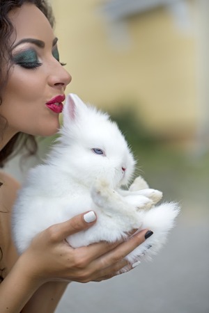 Closeup of pretty young brunette smiling lady with curly hair and bright makeup kissing and holding cute small fluffy white rabbit near face outdoor, vertical pictureの写真素材