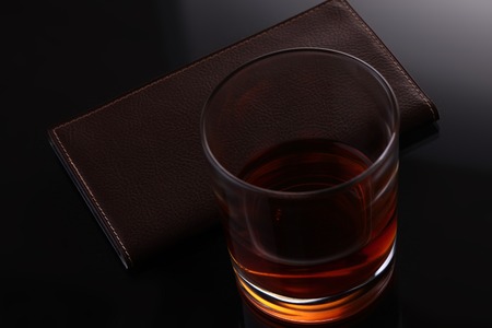 Glass of whiskey or other strong amber alcoholic beverage and leather brown purse on black reflecting table top studio background, horizontal pictureの写真素材