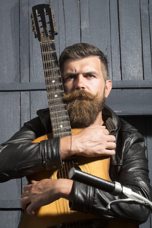 One serious unshaven musical man with beard and handlebar moustache holding acoustic string guitar with finger-board looking forward indoor on wooden wall black and white, vertical pictureの写真素材