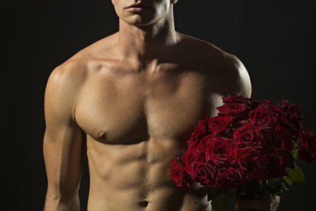 Attractive sexy undressed man with beautiful strong muscular body holding big fresh red rose flowers bouquet standing on dark grey background, horizontal pictureの写真素材