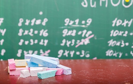 Blur texture background of school blackboard with written multiplication table and heap of colorful chalk blue pink violet, white yellow green colors lying on brown desk, horizontal pictureの写真素材