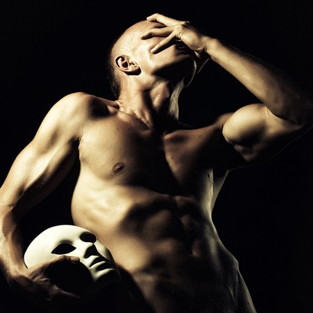 One young bare faceless man with sexual strong muscular attractive body with raised hand and beautiful chest holding one white mask standing on black studio background, square pictureの写真素材