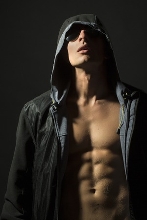 Young undressed guy with sexy strong muscular beautiful body in black leather jacket with hood and sunglasses standing on studio grey background, vertical pictureの写真素材