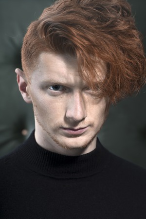 Portrait of handsome stylish young unshaven guy model with red hair standing in black jersey looking away indoor on studio background closeup, vertical pictureの写真素材