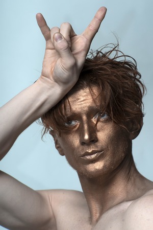 Portrait of young fashionable painted man model with bronzed bodyart on face and stylish hairdo holding hands in cool gesture looking away standing in studio on blue background, vertical pictureの写真素材