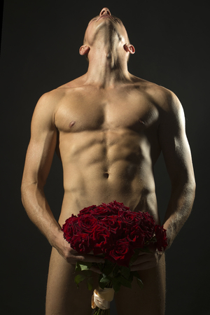 Handsome sexy undressed man with beautiful strong muscular body holding big fresh red rose flowers bouquet on genitals standing on grey background, vertical pictureの写真素材