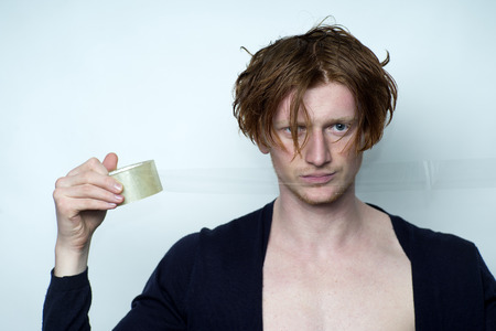 Portrait of sexy attractive young man model with red hair in black jersey with beautiful chest holding scotch tape looking forward standing in studio on white background copyspace, horizontal pictureの写真素材