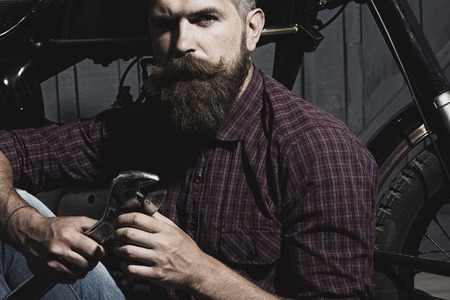 Young unshaven male biker in purple checkered shirt sitting near motorcycle in garage holding metallized iron wrench looking forward on workshop background, horizontal pictureの写真素材
