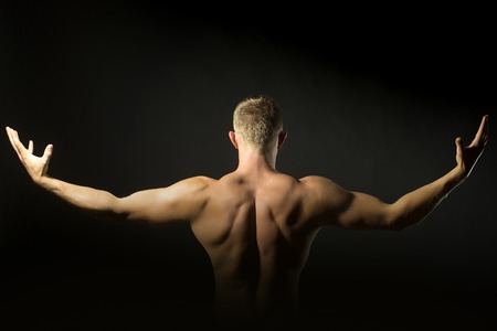 One young undressed man with sexual strong muscular attractive body with raised arms in sides and beautiful back standing on black studio background, horizontal pictureの写真素材