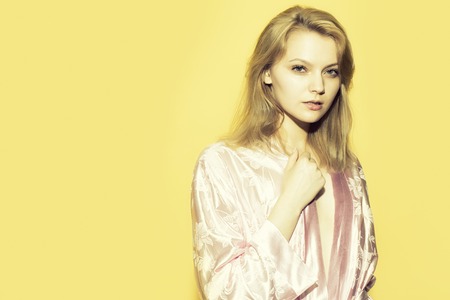 One beautiful young sexy pensive woman with blonde hair in satin pink dressing gown in soft flower pattern pastel colour standing on yellow studio background copyspace, horizontal pictureの写真素材