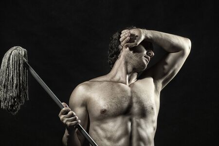 Handsome young sexy muscular athletic unshaven man cleaner with brunette curly hair and bare beautiful torso holding swab standing with raised hand on black studio background, horizontal pictureの写真素材