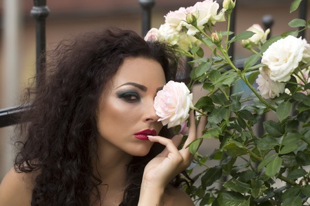 Pretty glamour sensual sexy young woman with curly brown long hair with fashion bright make-up emerald eyeshadow and red lips looking straight near white rose on blur background outdoor, horizontalの写真素材
