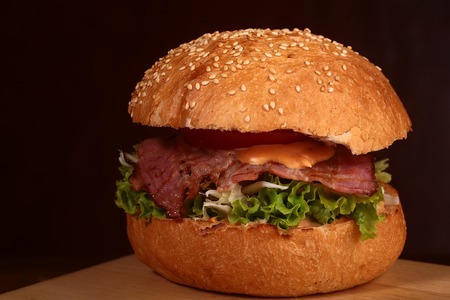 One big delicious fresh burger with green lettuce red tomato cheese cabbage bacon slice meat cutlet and white bread bun with sesame seeds on black background closeup, horizontal pictureの写真素材