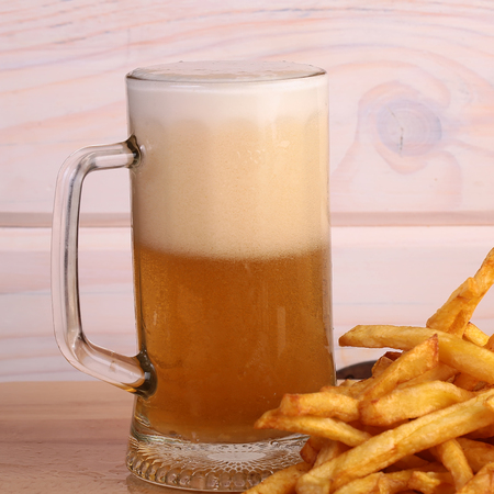 One big glass bocal of light cold delicious beer with white froth and tasty crispy yellow potato fasfood chips on wooden background closeup, square pictureの写真素材