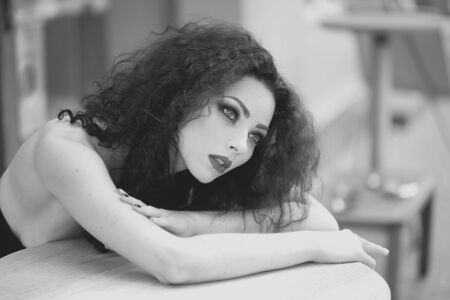 Charming cute young woman with professional fashion makeup curly hair healthy skin with bare shoulders looking away dreamy sitting at table black and white indoor on blur background, horizontalの写真素材