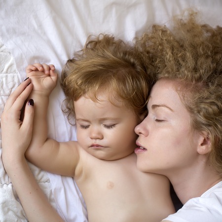 Closeup of tender beautiful loving mother with light blonde curly hair embracing small tiny cute male lovely baby sleeping indoor in bed with white linen lying close to each other, square pictureの写真素材