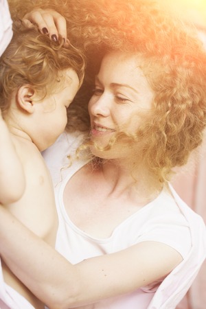 Closeup of smiling tender young loving mother with light blonde curly hair holding little tiny cute male lovely baby indoor in bed with white linen lying close to each other, horizontal pictureの写真素材