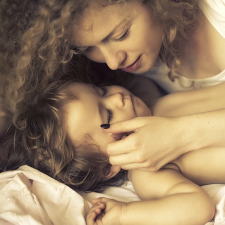 Closeup of young beautiful loving mother with light blonde curly hair sleeping with little tiny cute lovely son indoor in bed with white linen lying close to each other, square pictureの写真素材