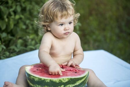 One small curious boy with blonde curly hair sitting on blue plaid holding half of fresh juicy green and red water melon with black seeds looking away on natural background, horizontal pictureの写真素材