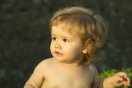 Closeup of little smiling interested cute baby boy with blonde curly hair round cheeks playful eyes standing without clothes outdoor lookling away on natural background, horizontal pictureの写真素材