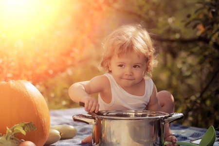 Closeup of one curious cook boy at picnic sitting with black ladle pot orange pumpkin and other vegetables playing with food sitting on blue checkered plaid on natural background, horizontal pictureの写真素材
