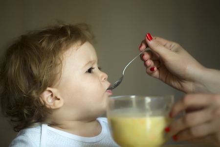 Nice little adorable toddler boy with hazel eyes and brown curly hair dressed in white t-shirt eating soup from glass bowl with spoon holding by mother on grey background indoor closeup, horizontalの写真素材