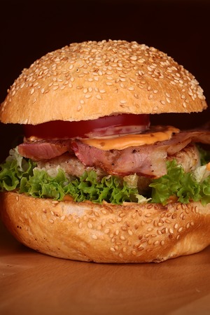 One appetizing big delicious fresh burger with green lettuce red tomato cheese cabbage bacon slice meat cutlet and white bread bun with sesame seeds on black background closeup, vertical pictureの写真素材