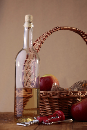 Composition of uncorked unlabel transparent glass bottle of aromatic sweet white wine wicker basket decorated with burlap apples and red corkscrew on brown wooden table, vertical photoの写真素材
