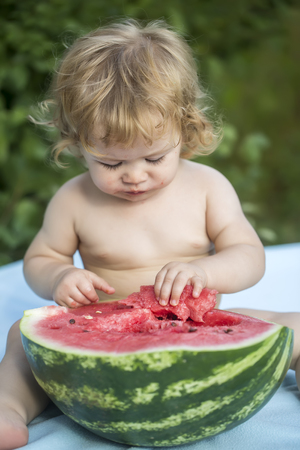 One little beautiful boy with blonde curly hair sitting on blue plaid eating with hands half of fresh juicy green and red water melon with black seeds looking down on natural background, vertical pictureの写真素材