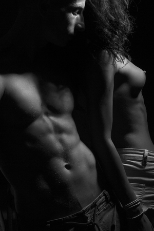 Half dressed sensual passionate pair of two people man with beautiful muscular body touching young woman with bare breast standing in studio close to each other black and white, vertical pictureの写真素材