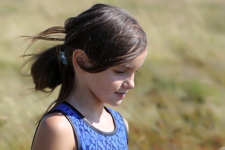 Portrait of pretty young brunette smiling girl with long hair in ponytail in blue lace dress looking down standing among yellow valley sunny day outdoor on natural blur background, horizontal photoの写真素材