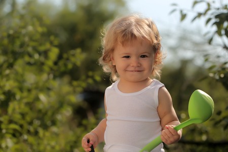 Portrait of happy smiling boy with blond curly hair in white underwear holding plastic ladle looking forward outdoor sunny day on natural background, horizontal pictureの写真素材