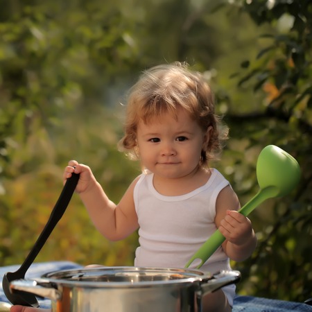Portrait of smiling boy with blonde curly hair in white underwear holding black and green kitchen utensil and cooking in big pot looking forward outdoor sunny day on natural background, square pictureの写真素材
