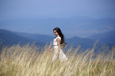 Cute smiling brunette girl in white lace summer dress walking in mountain valley among deep dry spikelet grass in spring sunny day outdoor on natural blue background, horizontal pictureの写真素材