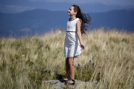 Little brunette girl in white lace summer dress standing in mountain valley with deep dry spikelet grass enjoying windy weather sunny day outdoor on natural blue background, horizontal pictureの写真素材